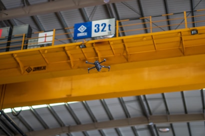 A drone is flying indoors near a large yellow overhead crane with a visible signage indicating 32t. The ceiling structure features metal beams and industrial lighting.