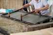 A person is engaged in traditional paper-making, using a wooden frame to scoop up a sheet of pulp from a water vat. The setting seems artisanal, with tools and materials neatly arranged around. The process appears to be taking place in a workshop-like environment.