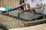 A person is engaged in traditional paper-making, using a wooden frame to scoop up a sheet of pulp from a water vat. The setting seems artisanal, with tools and materials neatly arranged around. The process appears to be taking place in a workshop-like environment.