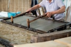 A person is engaged in traditional paper-making, using a wooden frame to scoop up a sheet of pulp from a water vat. The setting seems artisanal, with tools and materials neatly arranged around. The process appears to be taking place in a workshop-like environment.
