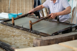 A person is engaged in traditional paper-making, using a wooden frame to scoop up a sheet of pulp from a water vat. The setting seems artisanal, with tools and materials neatly arranged around. The process appears to be taking place in a workshop-like environment.