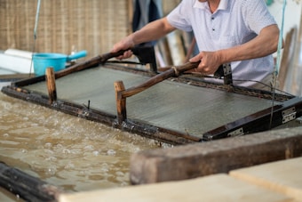 A person is engaged in traditional paper-making, using a wooden frame to scoop up a sheet of pulp from a water vat. The setting seems artisanal, with tools and materials neatly arranged around. The process appears to be taking place in a workshop-like environment.
