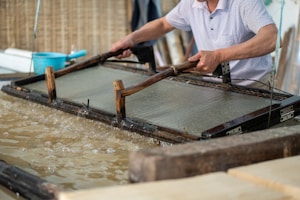 A person is engaged in traditional paper-making, using a wooden frame to scoop up a sheet of pulp from a water vat. The setting seems artisanal, with tools and materials neatly arranged around. The process appears to be taking place in a workshop-like environment.