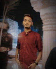 A young man stands confidently with hands in pockets, wearing a maroon shirt. The background shows blurry architectural elements and a palm tree, suggesting a night setting.
