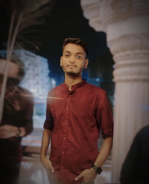 A young man stands confidently with hands in pockets, wearing a maroon shirt. The background shows blurry architectural elements and a palm tree, suggesting a night setting.