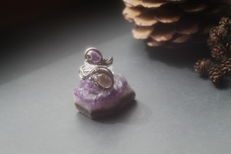 A silver ring with a purple gemstone sits atop a piece of amethyst crystal. The scene is softly lit, creating a serene and elegant atmosphere, with a pine cone placed in the background.
