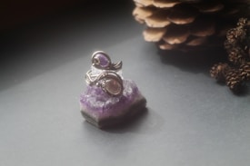 A silver ring with a purple gemstone sits atop a piece of amethyst crystal. The scene is softly lit, creating a serene and elegant atmosphere, with a pine cone placed in the background.
