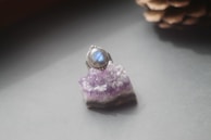 A silver ring with a bluish-purple gemstone is placed atop a purple crystal cluster. The background is softly blurred, with a pine cone partially visible in the top right corner, suggesting a natural or cozy setting.
