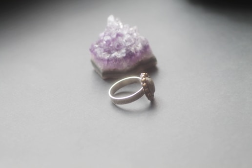 A silver ring with an ornate design is placed on a smooth, light gray surface. Behind it is a piece of raw amethyst crystal, displaying a vibrant purple color with a rough texture.