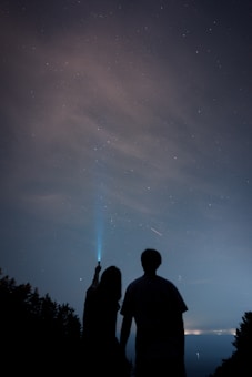 Silhouettes of two people stand against a night sky filled with stars. One person is holding up a light, pointing towards the sky. Trees outline the horizon, and the atmosphere conveys a sense of quiet wonder and exploration.