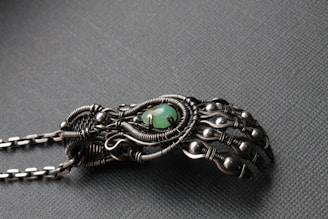 An intricately designed metallic hand sculpture featuring detailed wirework and spherical accents, with a central green stone. The craftsmanship highlights an organic but mechanical aesthetic with a matte finish.