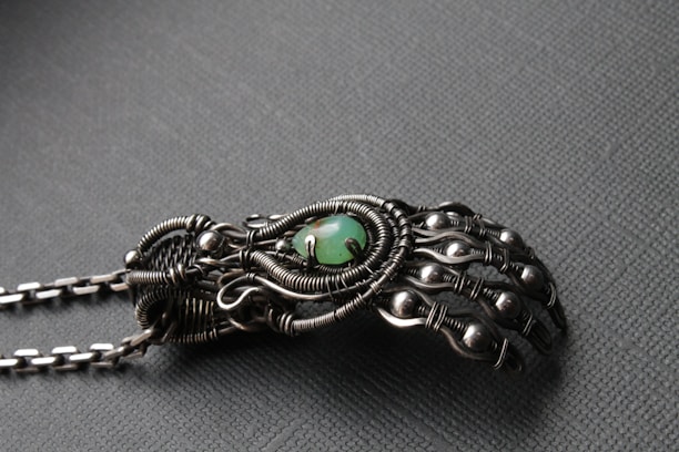 An intricately designed metallic hand sculpture featuring detailed wirework and spherical accents, with a central green stone. The craftsmanship highlights an organic but mechanical aesthetic with a matte finish.