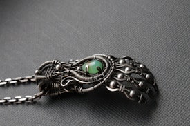 An intricately designed metallic hand sculpture featuring detailed wirework and spherical accents, with a central green stone. The craftsmanship highlights an organic but mechanical aesthetic with a matte finish.