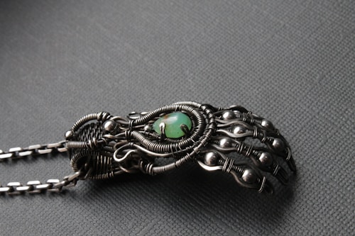 An intricately designed metallic hand sculpture featuring detailed wirework and spherical accents, with a central green stone. The craftsmanship highlights an organic but mechanical aesthetic with a matte finish.
