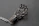A metallic sculpture resembling a hand, composed of intricate wirework and small spherical elements, is placed on a textured dark surface. The fingers are elongated and structured in a mechanical, futuristic design.