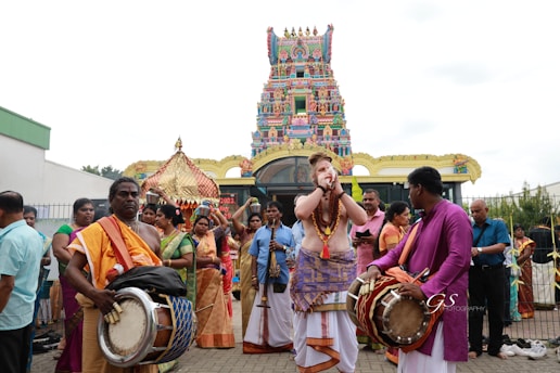 A group of people dressed in traditional clothing are engaged in a celebratory or religious procession in front of a colorful and intricately decorated temple. Several individuals are playing musical instruments such as drums and wind instruments, while others are observing or participating. The temple features vibrant patterns and statues on its façade.