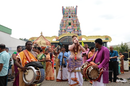A group of people dressed in traditional clothing are engaged in a celebratory or religious procession in front of a colorful and intricately decorated temple. Several individuals are playing musical instruments such as drums and wind instruments, while others are observing or participating. The temple features vibrant patterns and statues on its façade.
