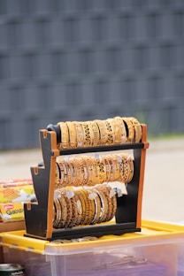 A stand displaying an assortment of intricately designed gold-colored bangles, some with detailed patterns, placed on a surface with various items in the background, including a plastic storage box and other objects.
