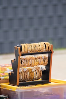 A stand displaying an assortment of intricately designed gold-colored bangles, some with detailed patterns, placed on a surface with various items in the background, including a plastic storage box and other objects.