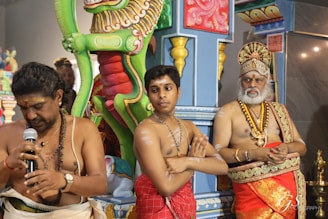 Three men dressed in traditional attire are standing in front of ornate, brightly colored temple decorations. One man holds a microphone, while another has his arms crossed. A third man is adorned with a decorative headdress and jewelry.