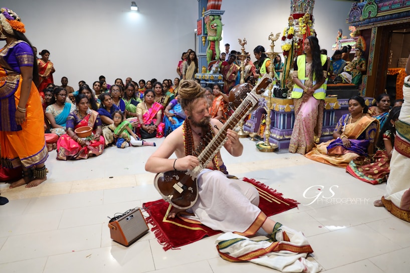 A musician is seated on a mat playing a traditional string instrument, surrounded by a large group of people. The setting appears to be a cultural or religious gathering with individuals dressed in brightly colored traditional attire. Intricate decorations and statues are visible in the background.
