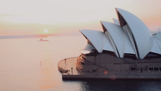 A view of the Sydney Opera House during sunset, with its iconic sail-like structures prominently on display. The sun is low on the horizon, casting a soft glow over the water in Sydney Harbour. There are few people walking along the promenade nearby, and a small boat can be seen in the distance.