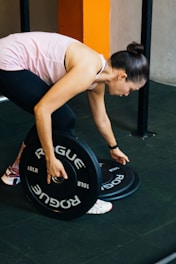 A person is bending down to lift or adjust a black barbell weight labeled 'ROGUE'. The person is wearing a pink tank top, black leggings, and white sneakers with colored details. The background includes a gym setting with a tile floor and a vertical orange accent wall.