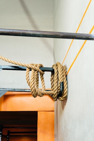Thick ropes are hanging from a metal bar against a textured wall. The ropes are coiled and draped over the bar, casting shadows on the light-colored wall. There are orange lines painted on the wall near the ropes, matching the orange rectangular structure on the lower part of the image.