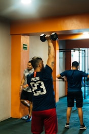 A group of people is engaged in a workout in a gym setting. One person in the foreground is lifting a dumbbell overhead with their back to the camera, wearing a jersey with 'Press 23' on it. Another person is visible in the background, leaning against an orange pillar. The environment is indoors with tiled flooring and a visible exercise apparatus in the background.