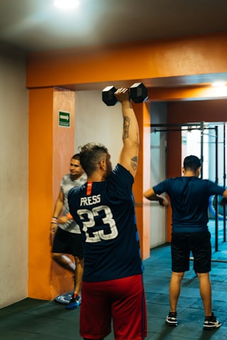 A group of people is engaged in a workout in a gym setting. One person in the foreground is lifting a dumbbell overhead with their back to the camera, wearing a jersey with 'Press 23' on it. Another person is visible in the background, leaning against an orange pillar. The environment is indoors with tiled flooring and a visible exercise apparatus in the background.