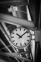 A large, round clock with a simple design and black hour and minute hands is mounted on a metal structure at a train station. The background features geometric patterns formed by beams and a corrugated metal ceiling, creating a sense of industrial architecture.
