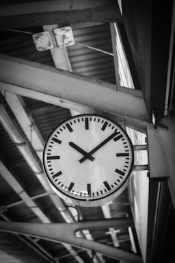 A large, round clock with a simple design and black hour and minute hands is mounted on a metal structure at a train station. The background features geometric patterns formed by beams and a corrugated metal ceiling, creating a sense of industrial architecture.