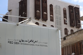 A modern event tent structure with the text 'Red Sea International Film Festival' is set against the backdrop of a traditional building featuring intricate wooden windows and decorative lattice panels.