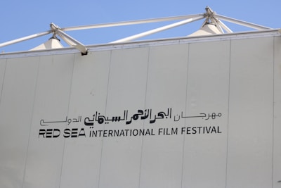 A large sign with bold writing in both Arabic and English letters. The words 'RED SEA INTERNATIONAL FILM FESTIVAL' are prominently displayed on a light-colored wall with a clear blue sky in the background and metal structural elements at the top.