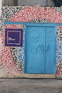 A vivid blue door with intricate designs is framed by walls covered in colorful Arabic calligraphy. The calligraphy features bold red, black, and blue letters on a white background. A sign with a deep purple background and the text 'Bait Alhodaif' is situated to the left of the door. The ground is made of stone tiles.