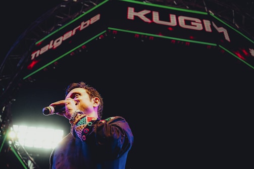 A person holding a microphone stands on stage under a large illuminated sign with a vibrant and colorful outfit. Bright lights shine in the background, creating an energetic atmosphere.