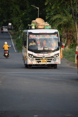 A colorful bus travels down a rural road, loaded with baskets and other items on its roof. Several passengers can be seen inside. The road is surrounded by dense greenery and a motorbike follows the bus in the distance.