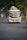 A colorful bus travels down a rural road, loaded with baskets and other items on its roof. Several passengers can be seen inside. The road is surrounded by dense greenery and a motorbike follows the bus in the distance.