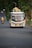 A colorful bus travels down a rural road, loaded with baskets and other items on its roof. Several passengers can be seen inside. The road is surrounded by dense greenery and a motorbike follows the bus in the distance.
