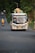 A colorful bus travels down a rural road, loaded with baskets and other items on its roof. Several passengers can be seen inside. The road is surrounded by dense greenery and a motorbike follows the bus in the distance.