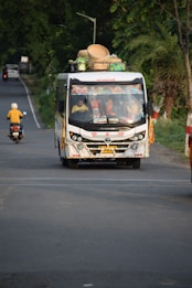 A colorful bus travels down a rural road, loaded with baskets and other items on its roof. Several passengers can be seen inside. The road is surrounded by dense greenery and a motorbike follows the bus in the distance.