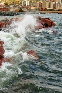 Waves crash against reddish-brown rocks along a shoreline with an urban cityscape in the background. Buildings rise in the distance, and the scene suggests a dynamic interaction between nature and urban development.