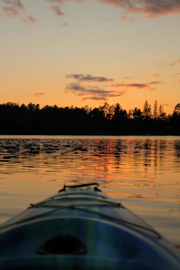 A serene kayak gliding through calm water at sunset, framed by lush trees.