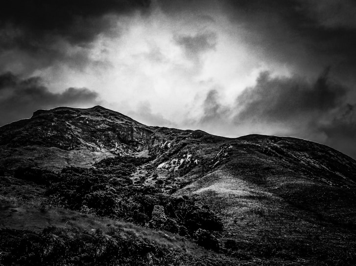 A dramatic landscape featuring a mountain with a rugged, uneven surface covered in patches of vegetation. The sky is filled with thick, dark, and swirling clouds that create an ominous atmosphere. The image is in black and white, adding to the overall stark and moody aesthetic.