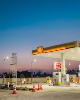A gas station at dusk with bright lights illuminating the area. The sky transitions from deep blue at the top to a lighter, warmer hue near the horizon. The station includes several fuel pumps under a shelter displaying the Shell logo, surrounded by orange traffic cones. There are signs and a small plant nearby, with street lamps providing additional lighting.