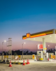 A gas station at dusk with bright lights illuminating the area. The sky transitions from deep blue at the top to a lighter, warmer hue near the horizon. The station includes several fuel pumps under a shelter displaying the Shell logo, surrounded by orange traffic cones. There are signs and a small plant nearby, with street lamps providing additional lighting.