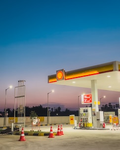 A gas station at dusk with bright lights illuminating the area. The sky transitions from deep blue at the top to a lighter, warmer hue near the horizon. The station includes several fuel pumps under a shelter displaying the Shell logo, surrounded by orange traffic cones. There are signs and a small plant nearby, with street lamps providing additional lighting.
