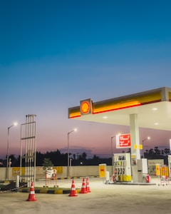 A gas station at dusk with bright lights illuminating the area. The sky transitions from deep blue at the top to a lighter, warmer hue near the horizon. The station includes several fuel pumps under a shelter displaying the Shell logo, surrounded by orange traffic cones. There are signs and a small plant nearby, with street lamps providing additional lighting.