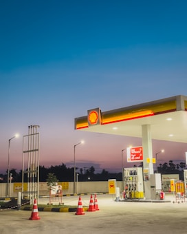 A gas station at dusk with bright lights illuminating the area. The sky transitions from deep blue at the top to a lighter, warmer hue near the horizon. The station includes several fuel pumps under a shelter displaying the Shell logo, surrounded by orange traffic cones. There are signs and a small plant nearby, with street lamps providing additional lighting.