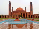A grand, ornate mosque featuring a large central dome flanked by two tall minarets, with intricate architectural details. The foreground includes a reflective pool with a central fountain and lush green landscaping. The structure is primarily made of reddish-brown brickwork with blue tile accents.